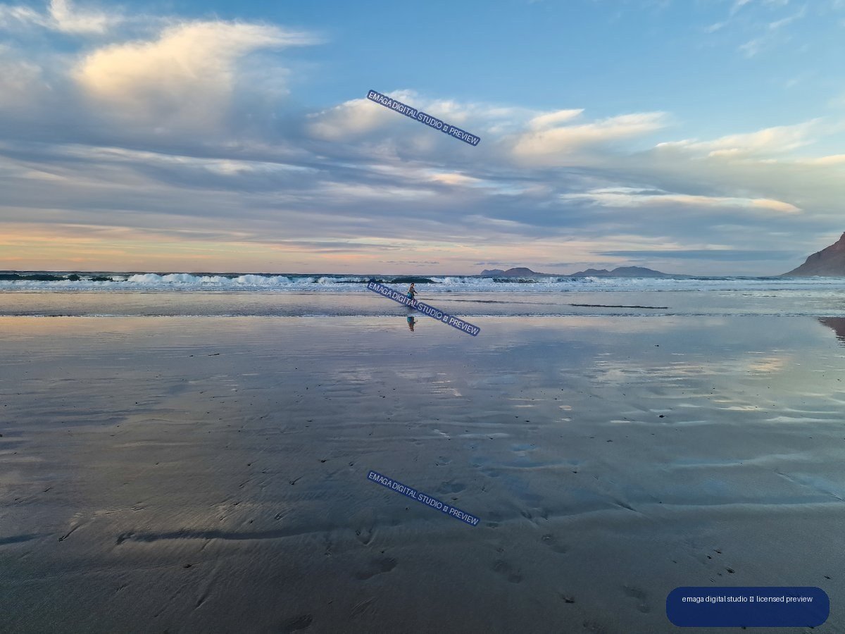 Wet reflective sand and shallow water at Famara Beach beneath a cloudy evening sky, with waves, a distant person, and mountains on the horizon.