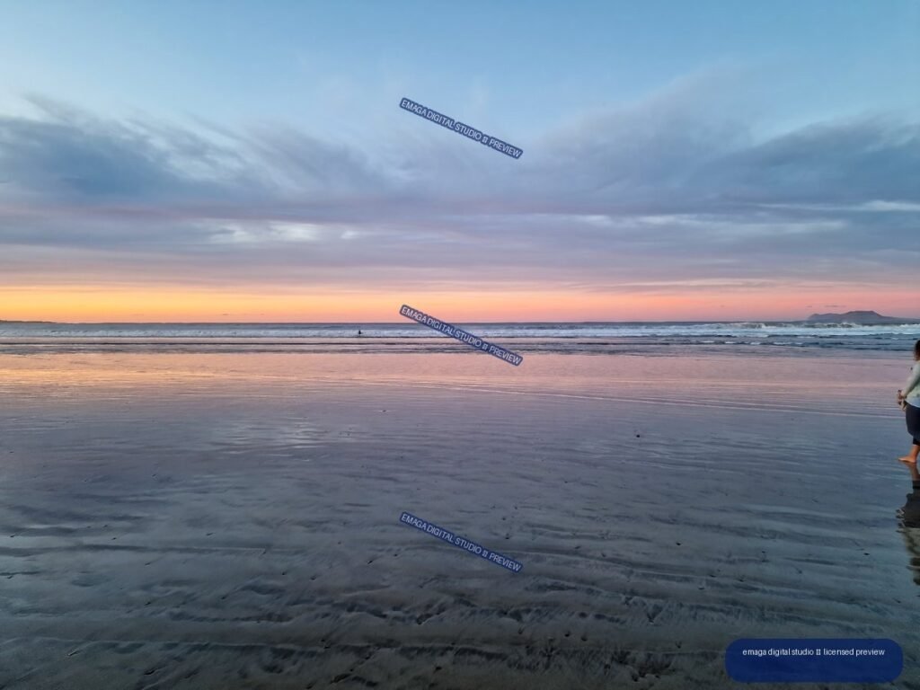 Wet reflective sand and gentle waves at Famara Beach beneath a pastel sunset sky, with distant hills on the horizon and people at the water's edge.
