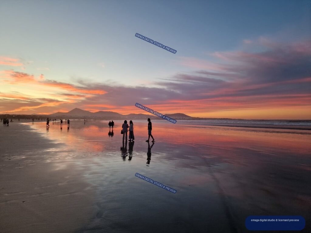 People walk across reflective wet sand at sunset on a broad beach, with volcanic mountains and colorful clouds on the horizon.