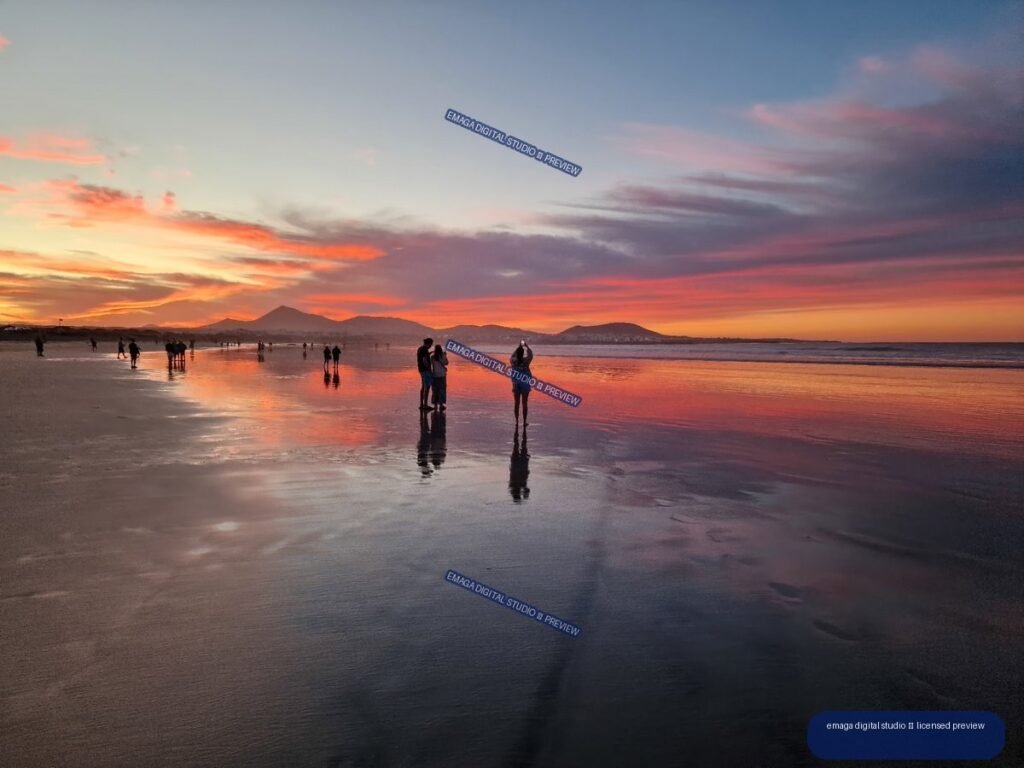 People on reflective wet sand at Famara Beach during a vivid sunset, with volcanic mountains on the horizon.
