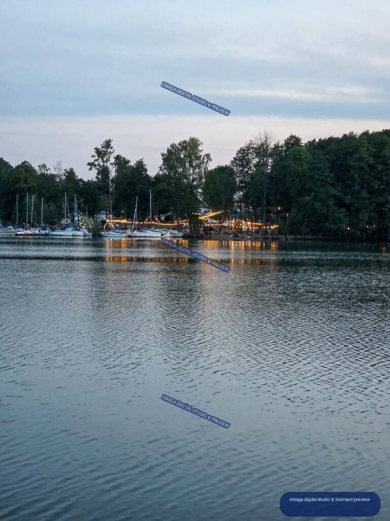Twilight view across rippling lake water toward sailboats, a wooded shore, and a marina glowing with warm string lights.