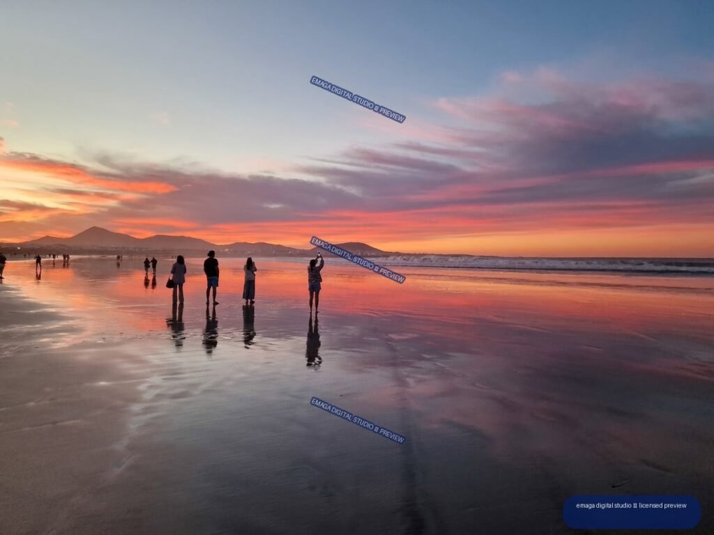 People and their reflections on wet sand at Famara Beach beneath a vivid pink and orange sunset, with volcanic hills on the horizon.