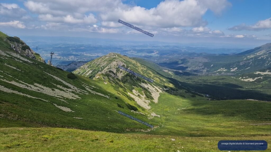 Panoramic alpine landscape with green Tatra slopes, rocky ridges, a valley, distant mountains, and cloudy blue sky.