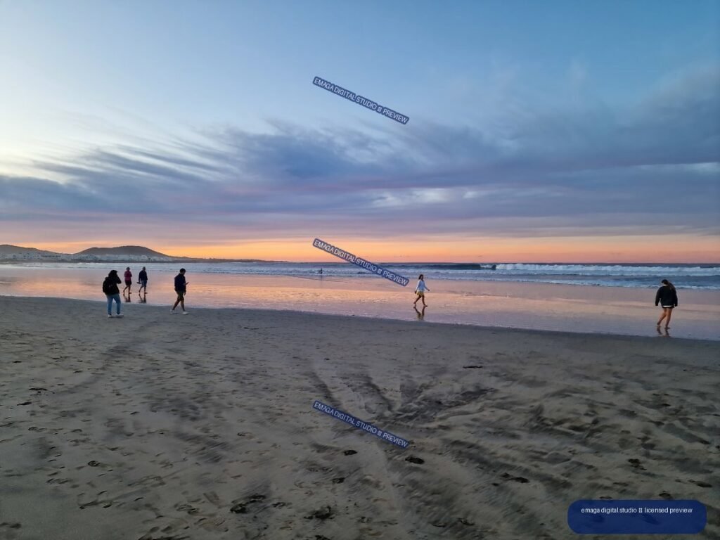 People walking along Famara Beach at sunset, with colorful sky reflections on wet sand and low hills in the distance.