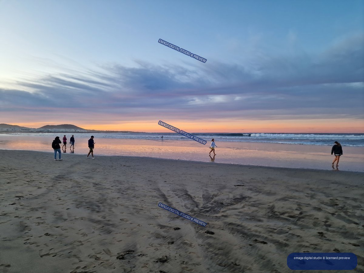 People walking along Famara Beach at sunset, with colorful sky reflections on wet sand and low hills in the distance.