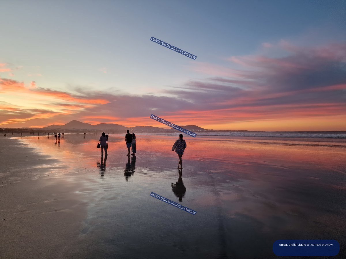 People and reflections on wet sand at Famara Beach beneath a colorful sunset, with mountains and ocean in the distance.