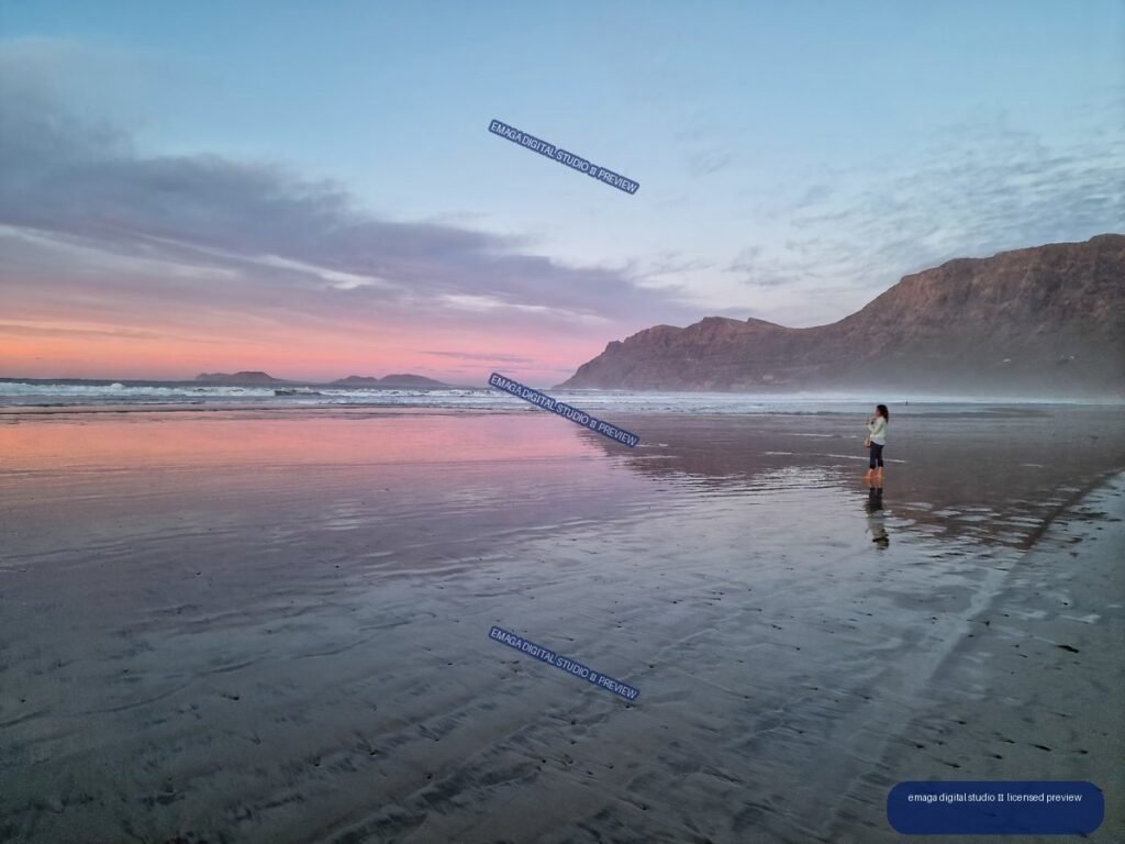 Wet reflective beach at dusk with pastel pink sky, waves, distant landforms, rugged cliffs, and one person near the shoreline.