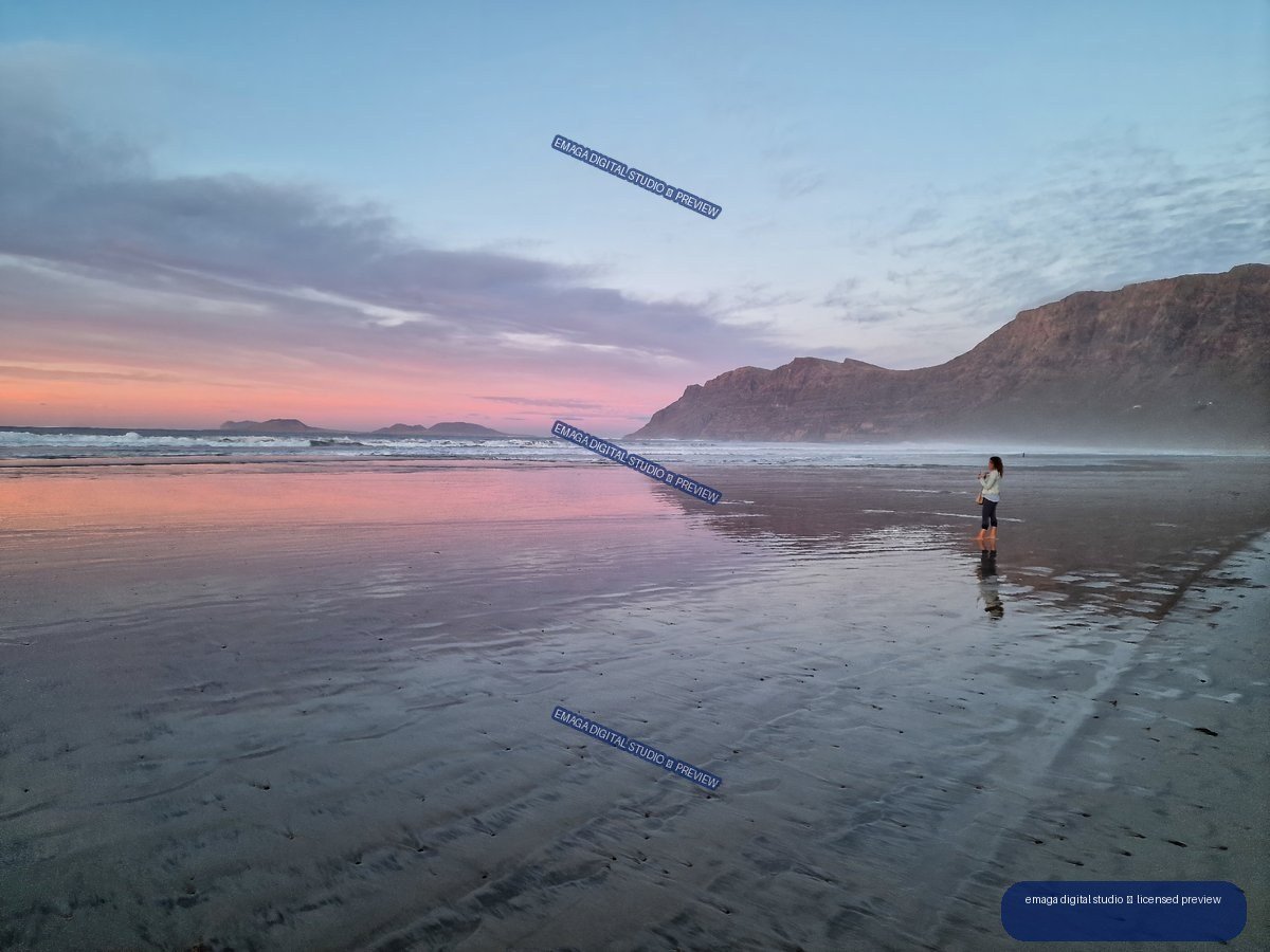 Wet reflective beach at dusk with pastel pink sky, waves, distant landforms, rugged cliffs, and one person near the shoreline.
