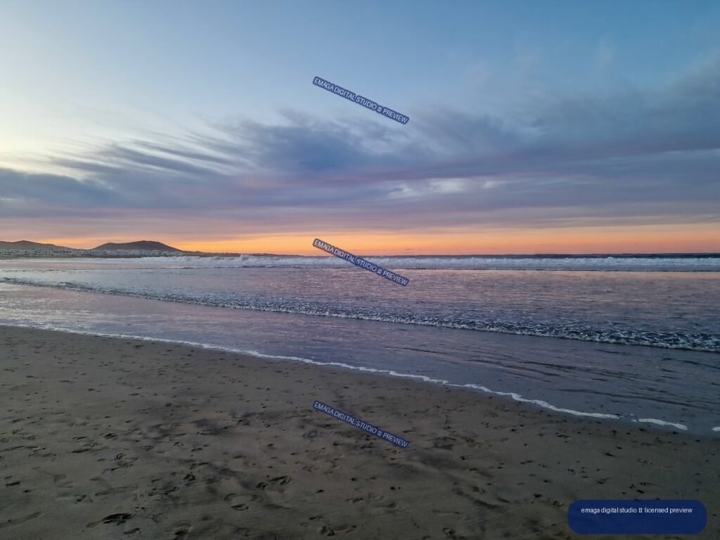 Sandy beach with reflective shallow water, gentle waves, distant hills, and a pink-orange sunset beneath layered blue-gray clouds.