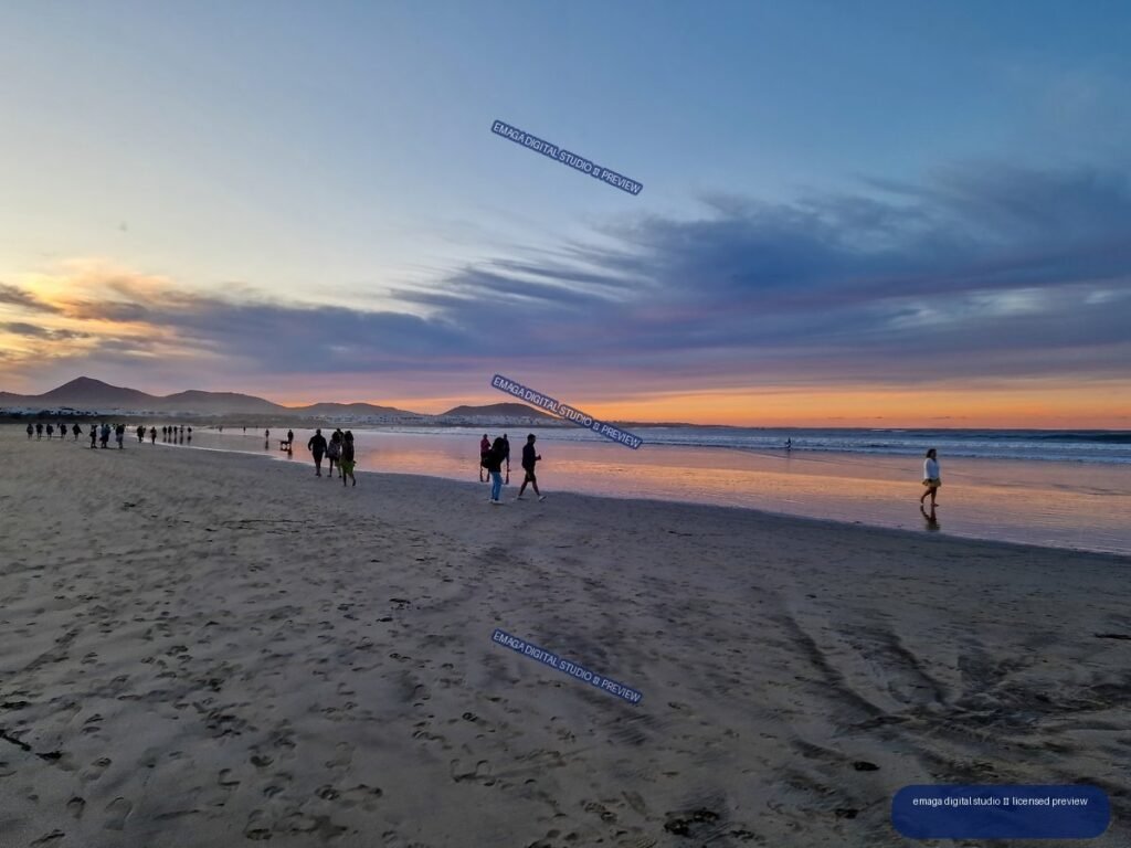 People walking along Famara Beach at sunset, with reflective wet sand, ocean waves, volcanic hills, and layered clouds.
