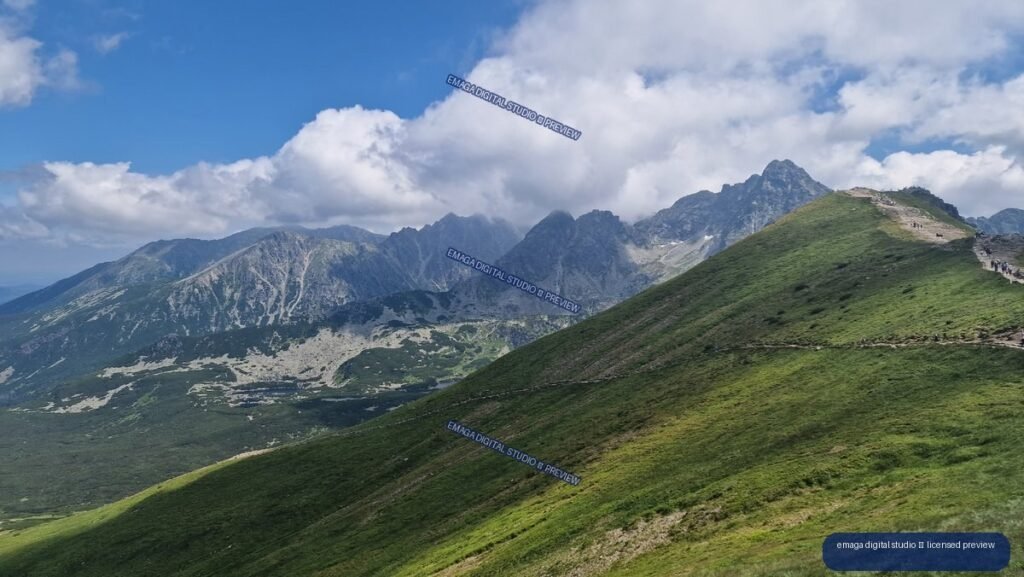Green alpine ridge with hikers overlooking rugged gray mountains beneath large white clouds and blue sky.