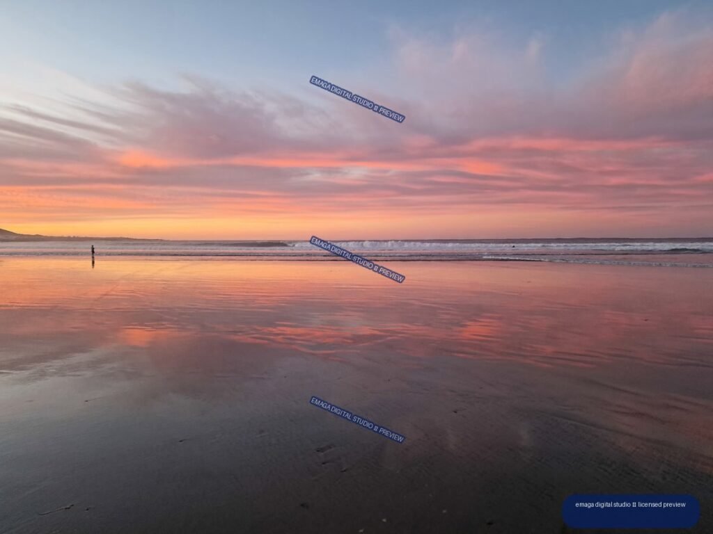 Pink and coral sunset reflected across wet sand beside a calm, wave-lined beach, with a small person near the left waterline.