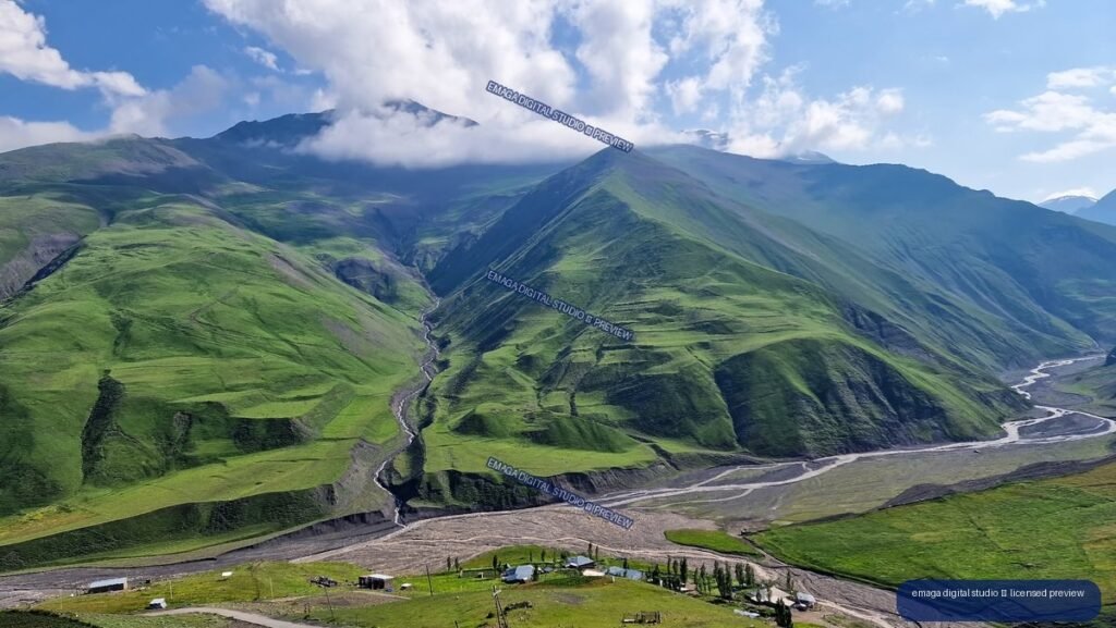 Panoramic view of green Caucasus mountains, a ravine, winding waterways, and small buildings beneath cloudy blue sky.