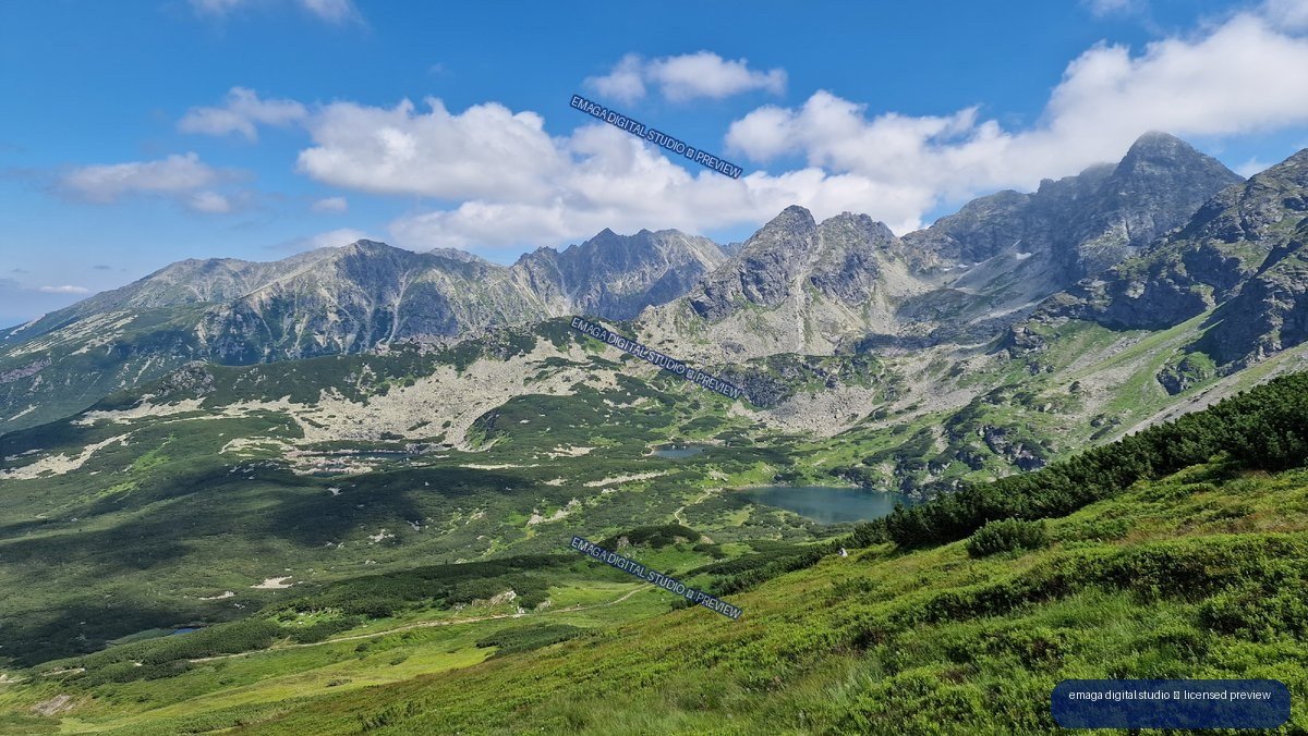 Panoramic view of rugged Tatra peaks, green slopes, alpine lakes, and a blue cloudy sky.