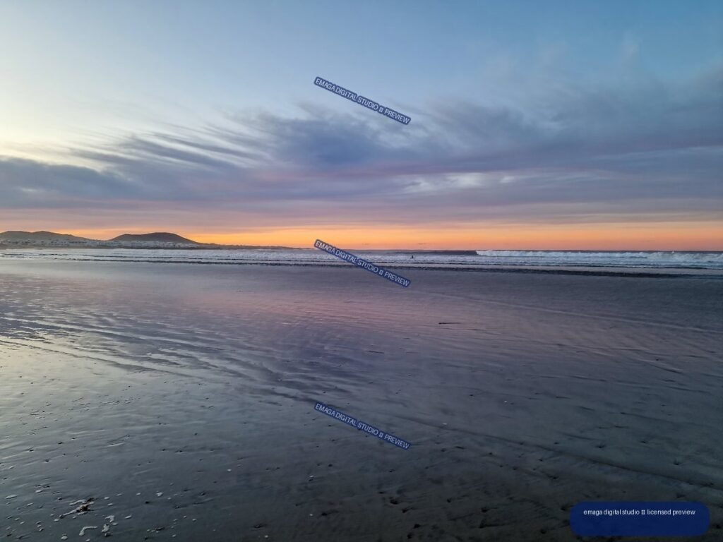 Wet reflective sand at Famara Beach mirrors a pastel sunset sky, with gentle waves and low hills on the horizon.