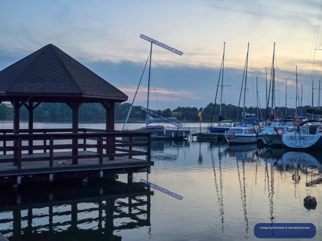 A lakeside marina at dusk with sailboats, a covered wooden pier, wooded shoreline, and reflections on calm water.