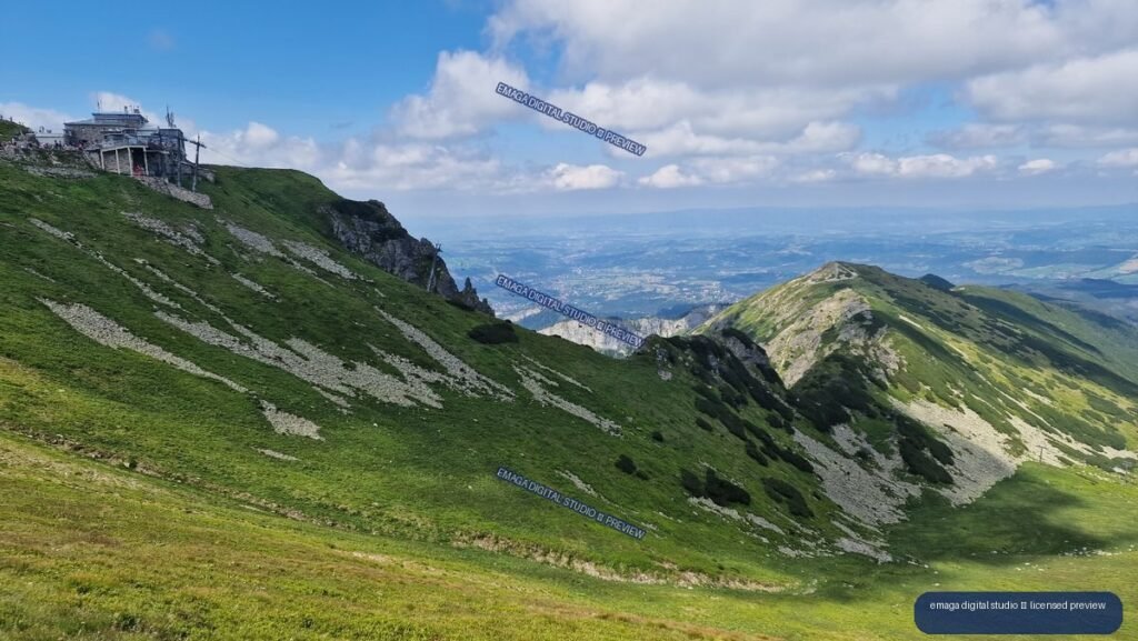Green rocky alpine slopes and a rugged ridge overlook distant valleys beneath a blue, cloud-filled sky; a mountain station appears at upper left.