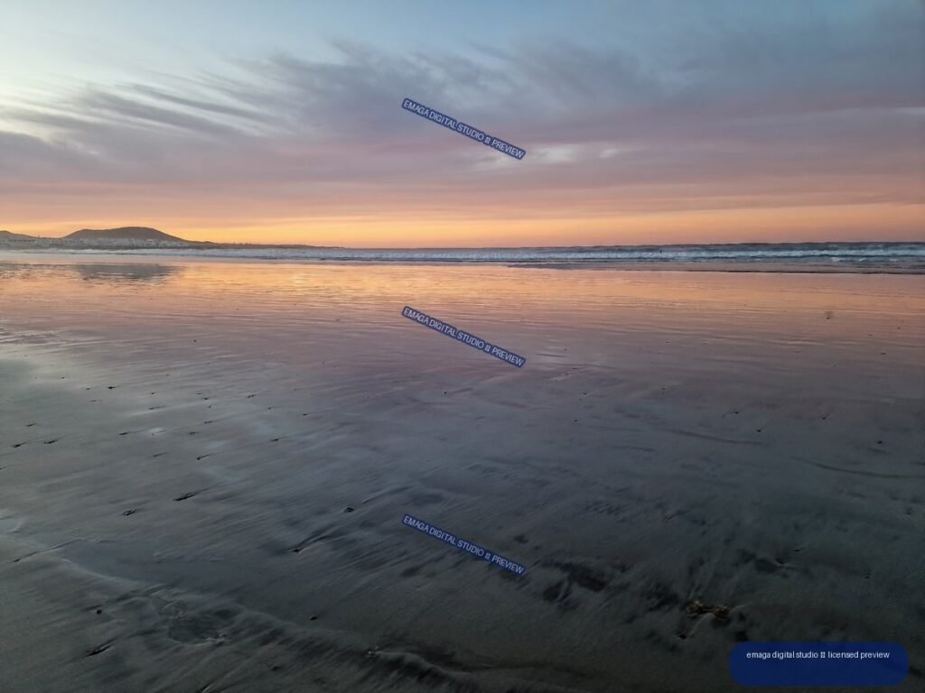 Wet reflective sand at Famara Beach mirrors a lavender and orange sunset sky, with gentle waves and a low headland on the horizon.