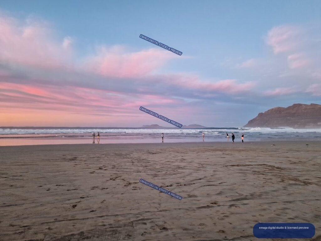 Wide view of Famara Beach at sunset with pink clouds, reflective wet sand, people near the waves, and a rugged headland on the right.