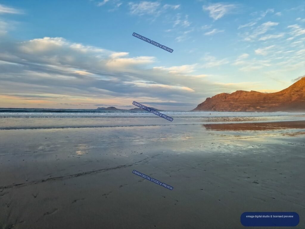Wet reflective beach at sunset with waves, distant islands, and a rugged reddish headland beneath a cloud-filled sky.