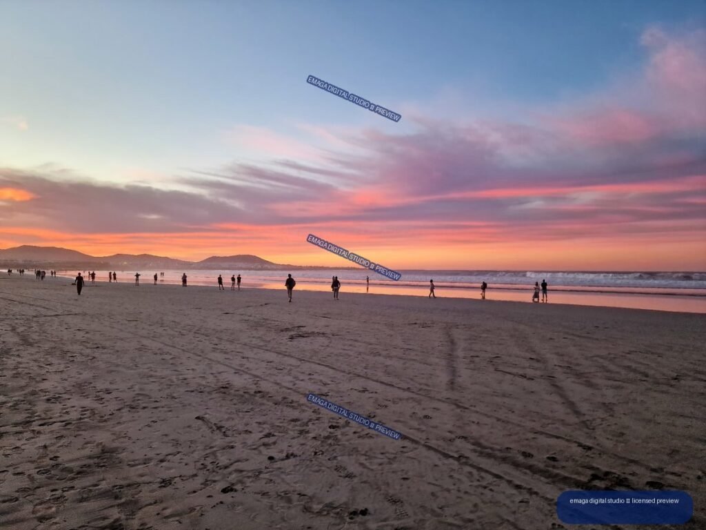 Wide sandy beach with silhouetted people, reflective shoreline, distant hills, and a pink-orange sunset sky.