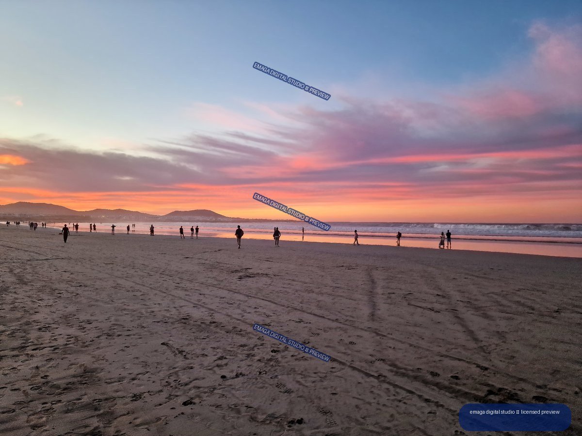 Wide sandy beach with silhouetted people, reflective shoreline, distant hills, and a pink-orange sunset sky.