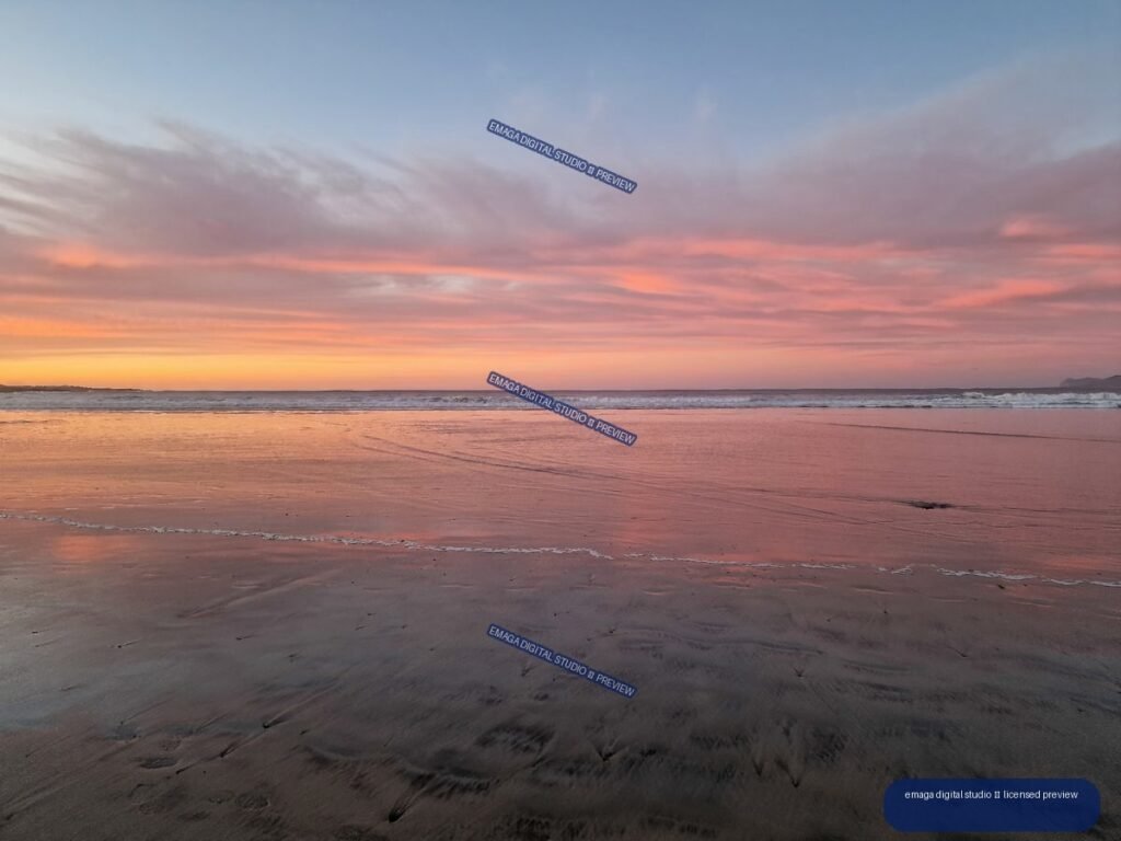 Pastel sunset reflected on wet sand at Famara Beach, with calm waves and distant shoreline.