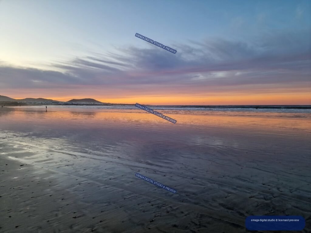 Wet sand reflecting a pink and orange sunset at Famara Beach, with distant hills, waves, and a lone figure.
