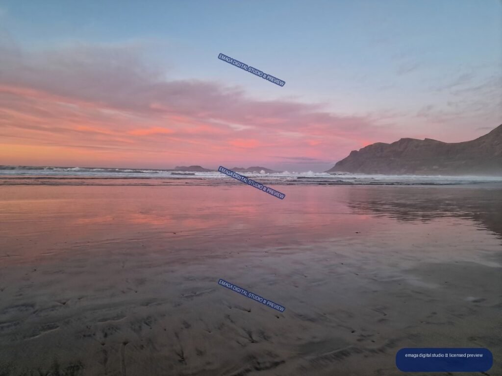 Wet sand reflecting a pink sunset at Famara Beach, with waves, distant landforms, and a rugged mountain headland.