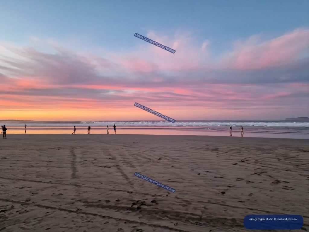 Wide beach at sunset with pink clouds, reflective shoreline, ocean waves, distant walkers, and textured wet sand.