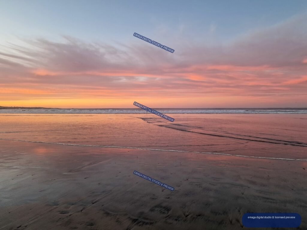 Reflective wet sand and shallow waves beneath a pink and orange sunset sky at a beach.