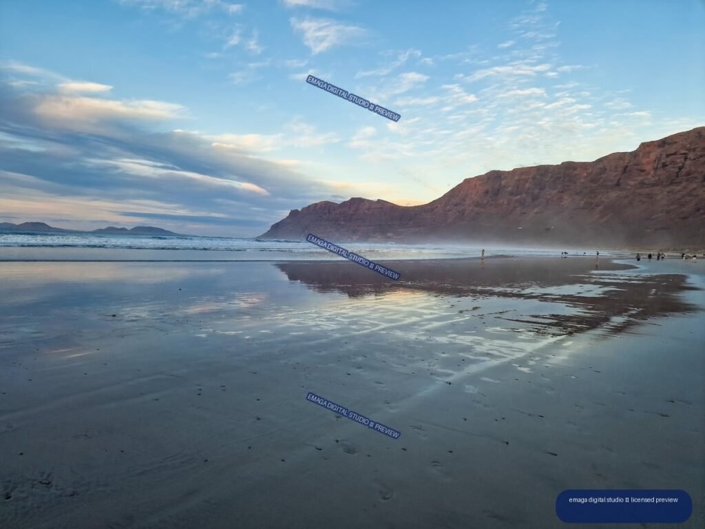 Wet reflective beach beneath reddish cliffs, with waves, clouds, and distant people at dusk.