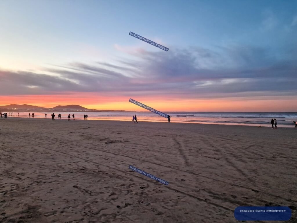 Wide view of Famara Beach at sunset with colorful reflections, distant hills, waves, clouds, and scattered people.