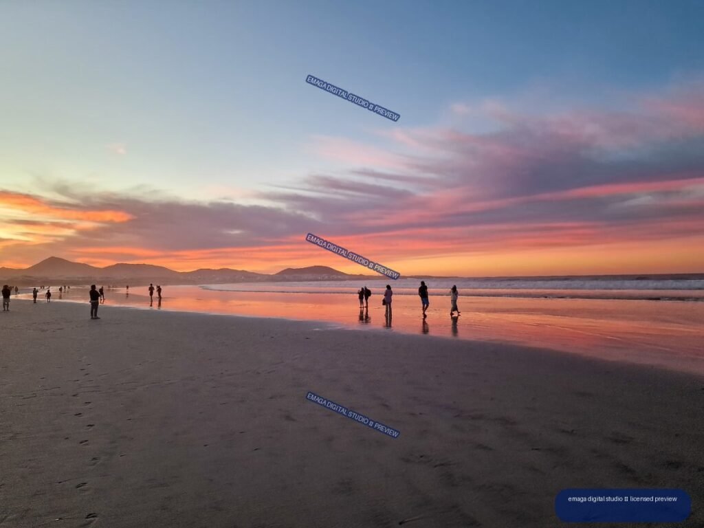 Sunset over a wide beach with colorful cloud reflections on wet sand, people along the shore, waves, and distant volcanic mountains.