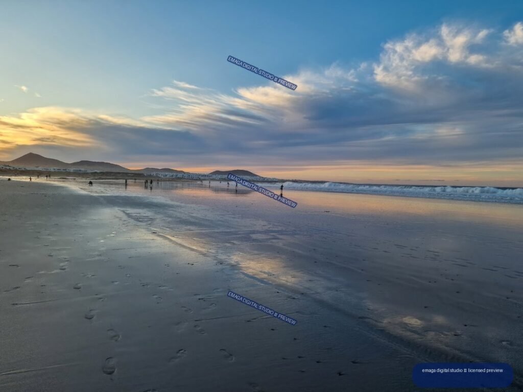 Wet reflective sand and gentle waves at Famara Beach beneath a cloud-filled sunset sky, with hills and distant buildings on the horizon.
