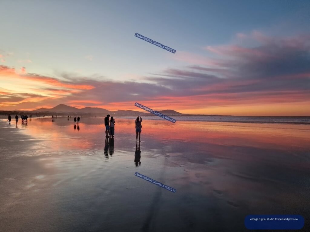 People walking across reflective wet sand at sunset, with mountains and colorful clouds along the horizon.
