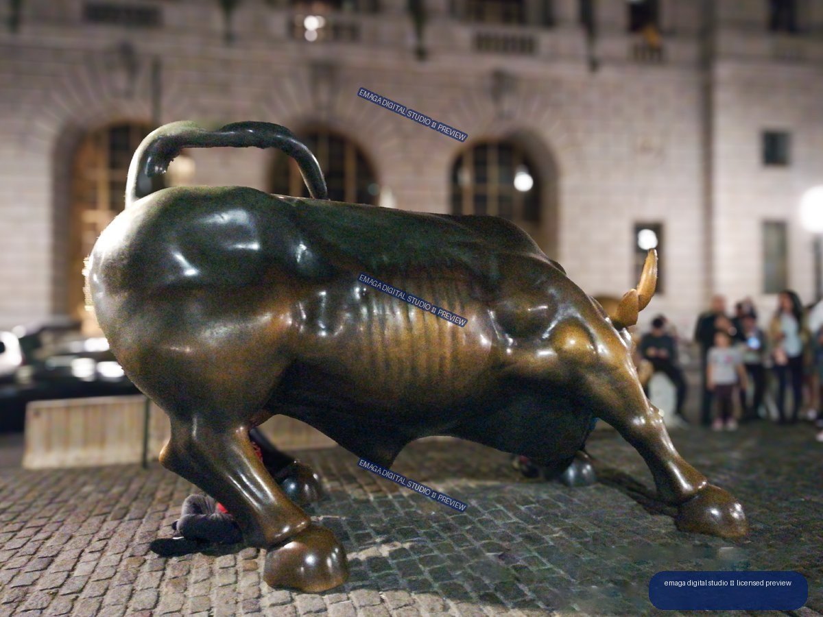 Large bronze charging bull sculpture on a cobblestone plaza at night, with visitors and an illuminated building behind it.