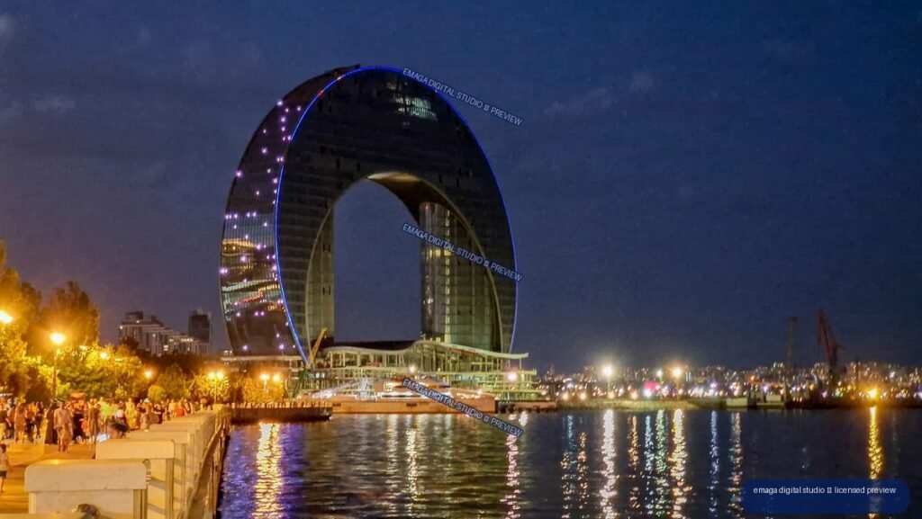 Nighttime waterfront with a large blue-lit elliptical arch, promenade, boats, city lights, and reflections on the water.