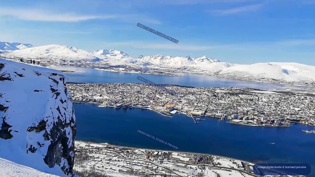 Aerial panorama of a snow-covered coastal city beside blue water, surrounded by rugged white mountains.