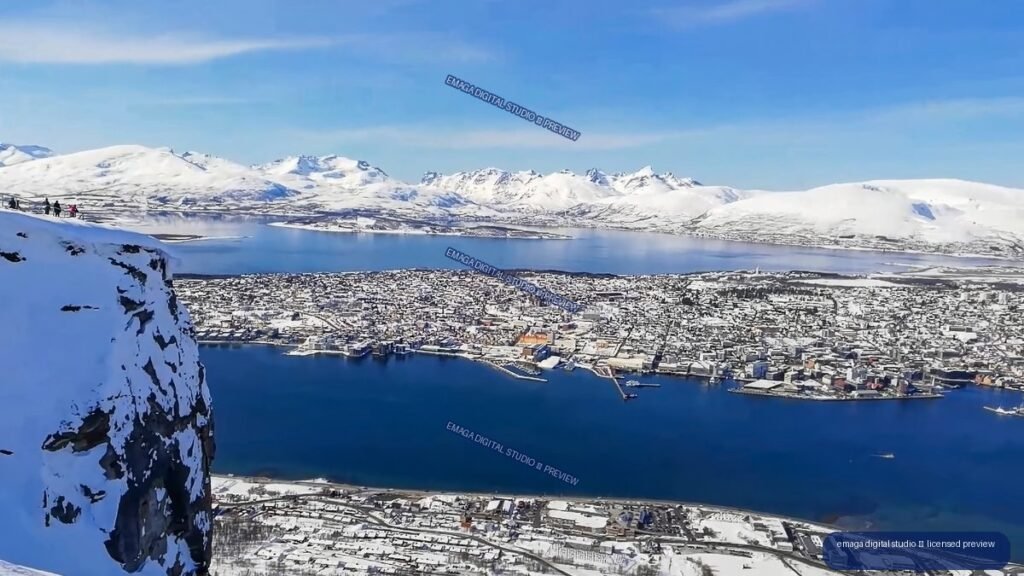 Elevated view of a snowy coastal city, blue water, mountains, and a snow-covered cliff in the foreground.