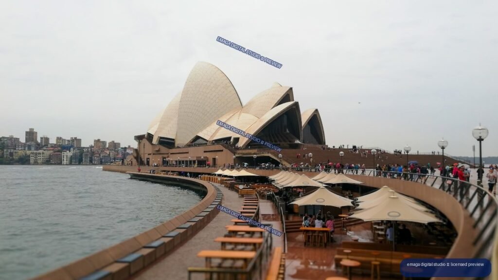 Sydney Opera House beside a curving harbor promenade with outdoor seating, visitors, and city buildings across the water.