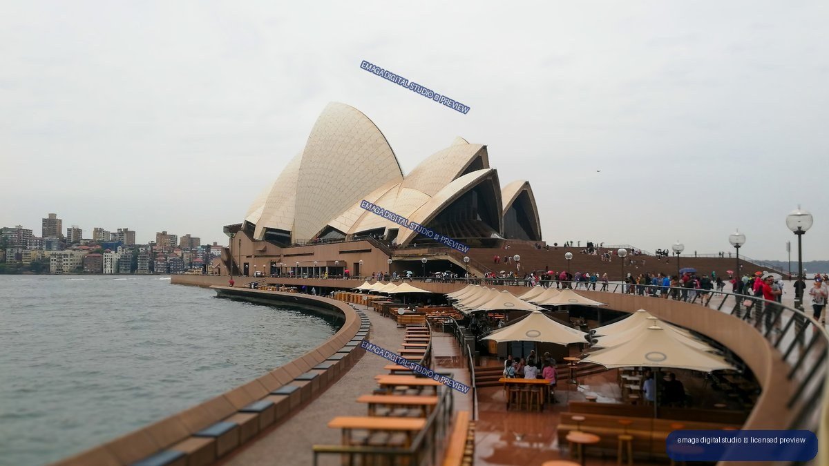 Sydney Opera House beside a curving harbor promenade with outdoor seating, visitors, and city buildings across the water.